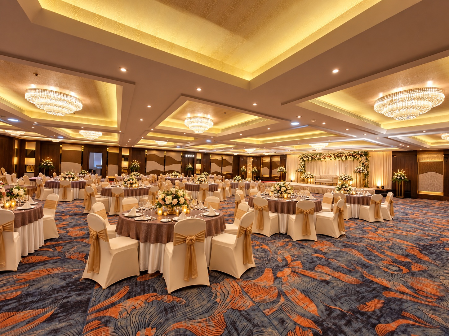 Décor concept at Veloria Grand Rajajinagar: taupe round tables, gold-sash chairs and crystal chandeliers facing a floral stage